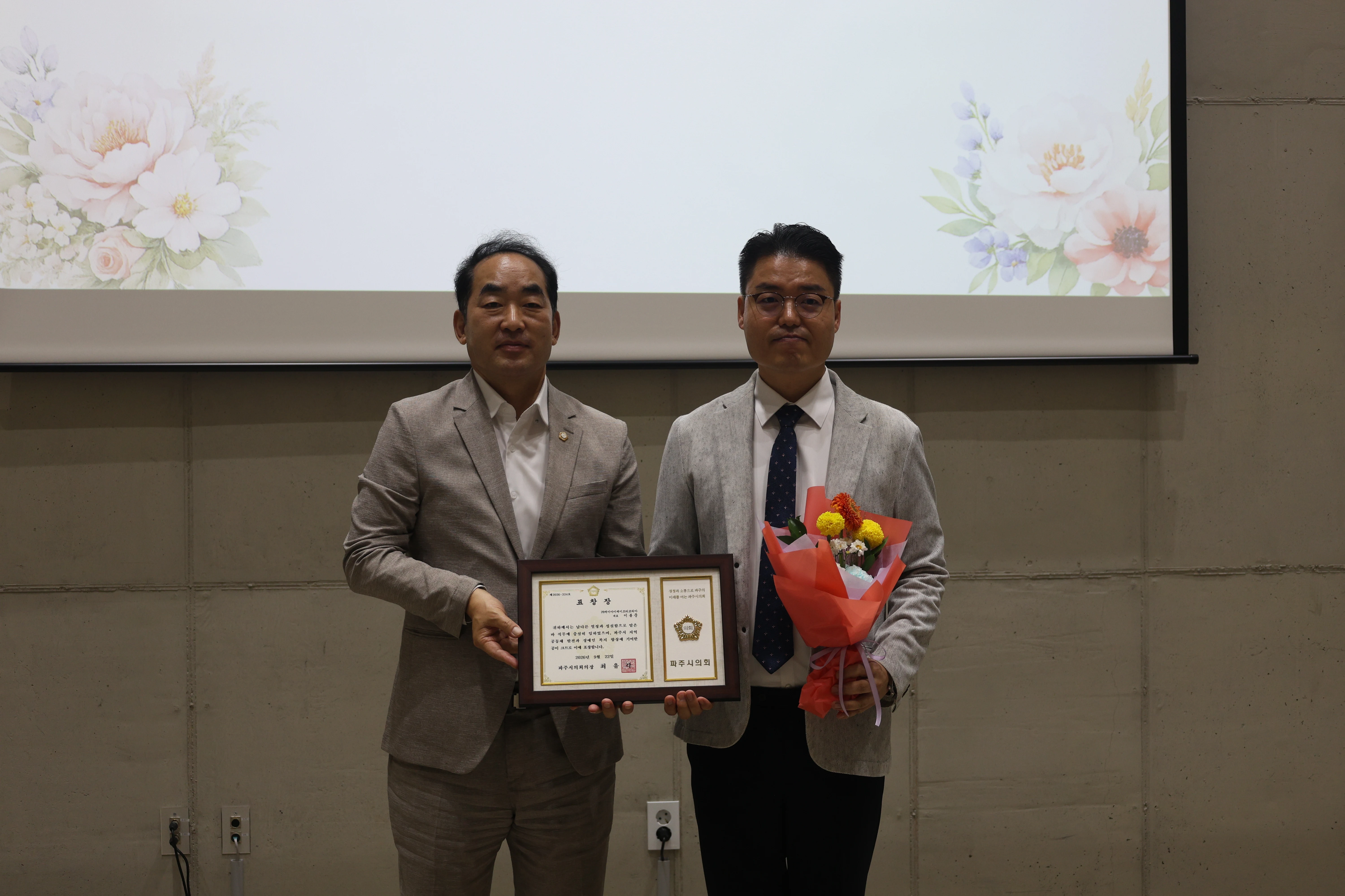 회색 재킷을 입은 두 남성이 행사 화면 앞에 서 있고, 오른쪽의 이봉준 창업자가 꽃다발을 든 채 파주시의회 의장상 표창패를 함께 들고 있다.